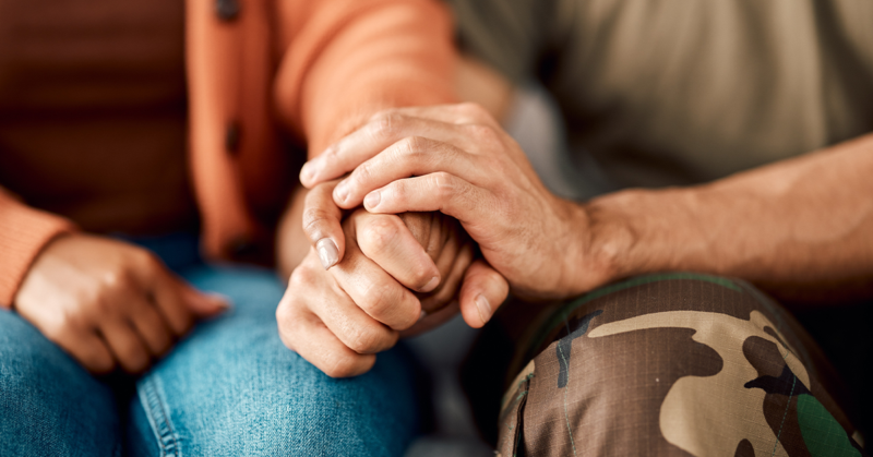 A close-up of two people sitting side by side, one in a brown sweater holding hands with another in camo pants.
