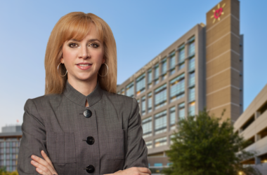 UMC new CEO Maria Zampini, confidently stands in front of a modern office building against a clear blue sky, conveying professionalism.