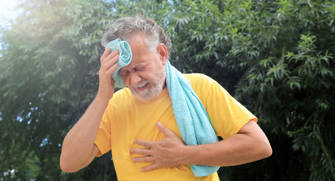 Elderly man in a yellow shirt wipes sweat from his brow with a towel. He appears distressed, clutching his chest, against a backdrop of green trees.