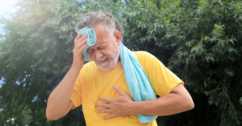 Elderly man in a yellow shirt wipes sweat from his brow with a towel. He appears distressed, clutching his chest, against a backdrop of green trees.