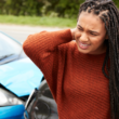A woman stands beside a car with a dented front, rubbing her neck in discomfort, representing trauma awareness.