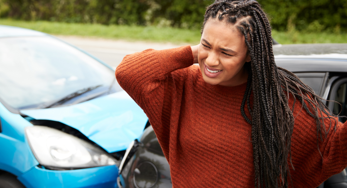 A woman stands beside a car with a dented front, rubbing her neck in discomfort, representing trauma awareness.