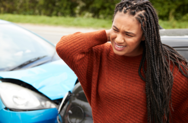 A woman stands beside a car with a dented front, rubbing her neck in discomfort, representing trauma awareness.