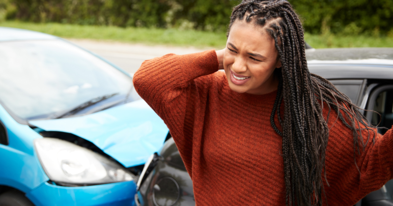 A woman stands beside a car with a dented front, rubbing her neck in discomfort, representing trauma awareness.
