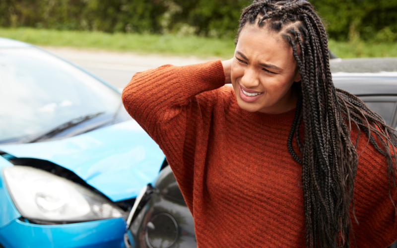 A woman stands beside a car with a dented front, rubbing her neck in discomfort, representing trauma awareness.