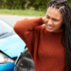 A woman stands beside a car with a dented front, rubbing her neck in discomfort, representing trauma awareness.