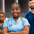 A confident young nurse with a stethoscope smiles in the foreground. Behind her, two medical professionals stand, conveying teamwork in a healthcare setting.