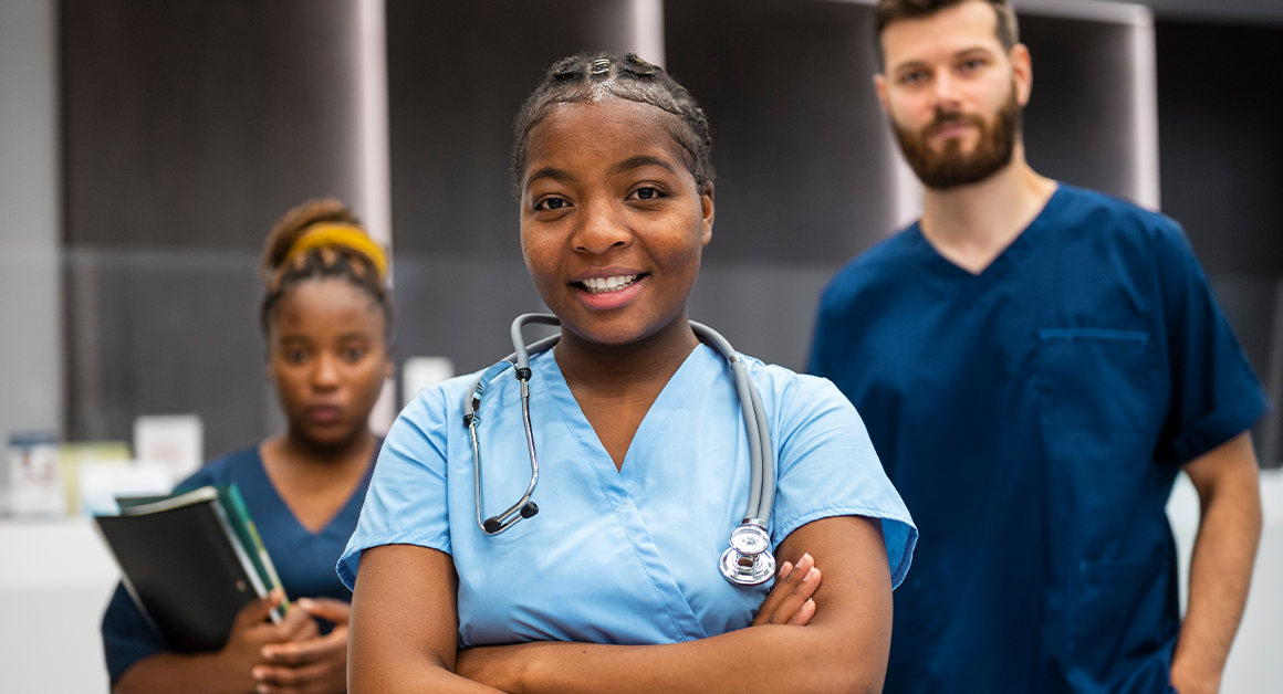A confident young nurse with a stethoscope smiles in the foreground. Behind her, two medical professionals stand, conveying teamwork in a healthcare setting.