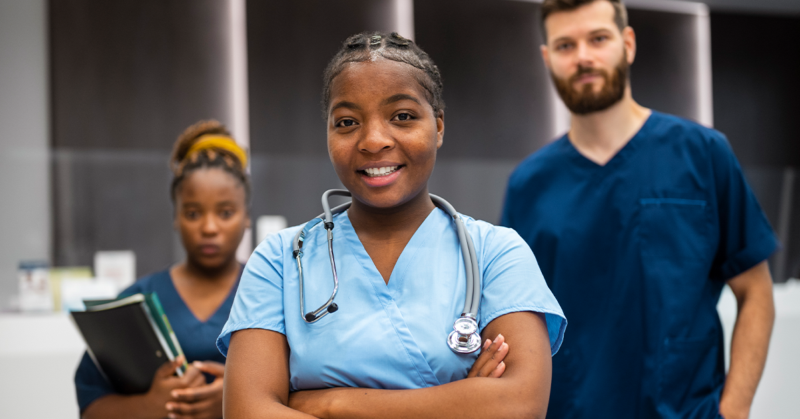 A confident young nurse with a stethoscope smiles in the foreground. Behind her, two medical professionals stand, conveying teamwork in a healthcare setting.