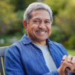Smiling older man with gray hair sits outdoors on a wooden chair, wearing a blue shirt. Lush, blurry garden background suggests a peaceful atmosphere.