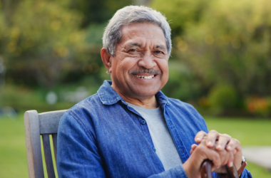 Smiling older man with gray hair sits outdoors on a wooden chair, wearing a blue shirt. Lush, blurry garden background suggests a peaceful atmosphere.