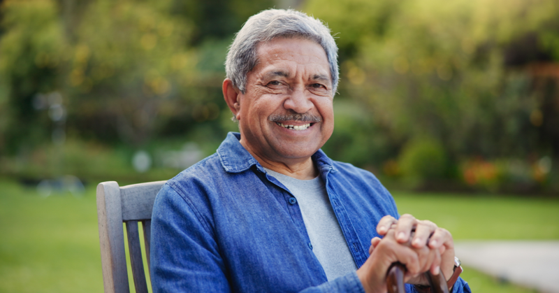 Smiling older man with gray hair sits outdoors on a wooden chair, wearing a blue shirt. Lush, blurry garden background suggests a peaceful atmosphere.