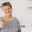 Elderly woman smiling and giving a thumbs-up in a bright kitchen. She wears glasses and a checkered blouse, exuding warmth and positivity.