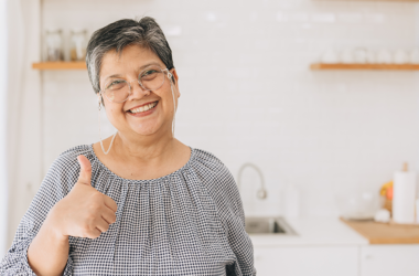 Elderly woman smiling and giving a thumbs-up in a bright kitchen. She wears glasses and a checkered blouse, exuding warmth and positivity.