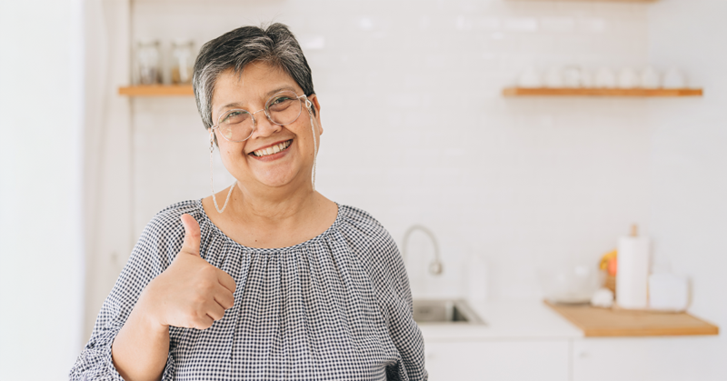 Elderly woman smiling and giving a thumbs-up in a bright kitchen. She wears glasses and a checkered blouse, exuding warmth and positivity.