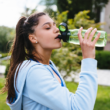 Woman in a blue hoodie drinks from a green water bottle outdoors. She's in a lush park setting, taking a refreshing break, conveying relaxation.