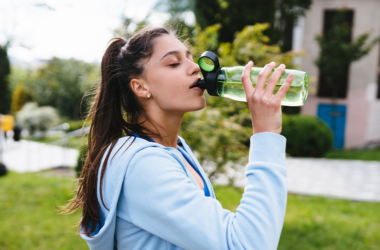 Woman in a blue hoodie drinks from a green water bottle outdoors. She's in a lush park setting, taking a refreshing break, conveying relaxation.