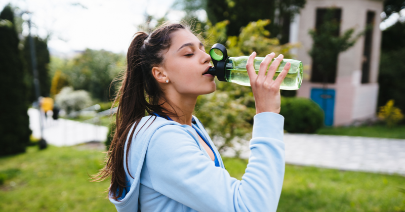 Woman in a blue hoodie drinks from a green water bottle outdoors. She's in a lush park setting, taking a refreshing break, conveying relaxation.