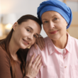 A loving portrait of two women; one with a blue headscarf, the other resting her head on her shoulder. They are indoors, representing National Cancer Survivors Day.