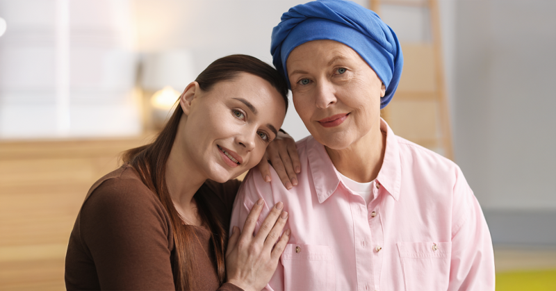 A loving portrait of two women; one with a blue headscarf, the other resting her head on her shoulder. They are indoors, representing National Cancer Survivors Day.