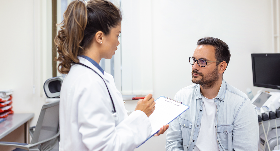 A doctor in a white coat holds a clipboard, attentively conversing with a seated man in a light shirt. The scene is a calm, clinical exam room.