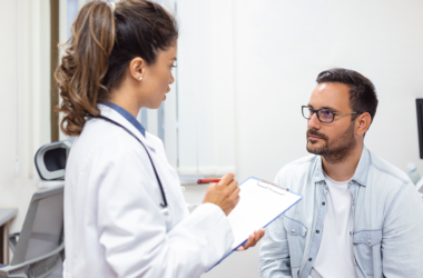A doctor in a white coat holds a clipboard, attentively conversing with a seated man in a light shirt. The scene is a calm, clinical exam room.