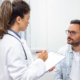 A doctor in a white coat holds a clipboard, attentively conversing with a seated man in a light shirt. The scene is a calm, clinical exam room.