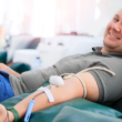 A man smiling while donating blood, reclined on a chair. A medical tube is connected to his arm, representing World Blood Donor Day.