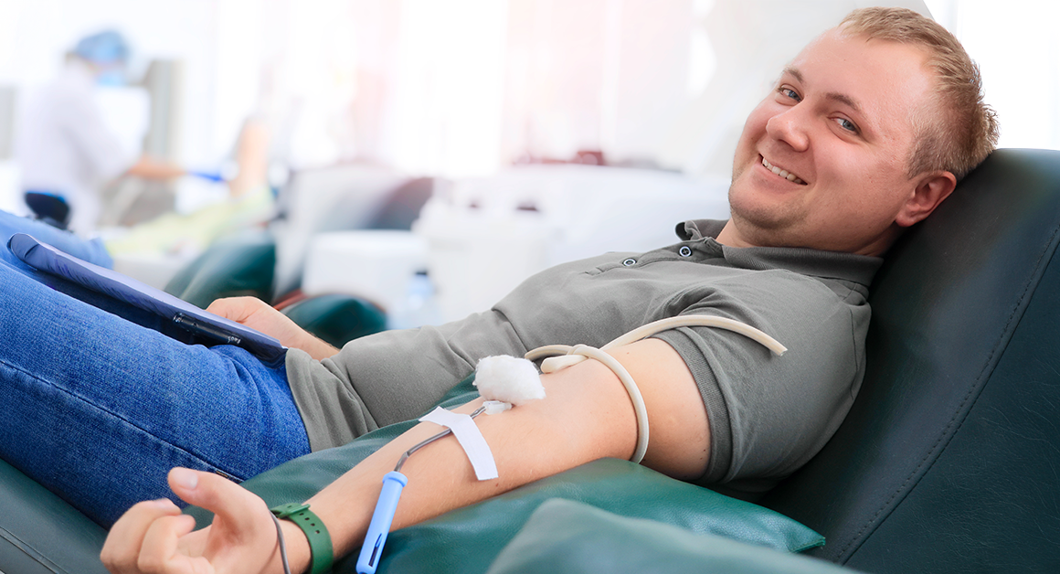 A man smiling while donating blood, reclined on a chair. A medical tube is connected to his arm, representing World Blood Donor Day.