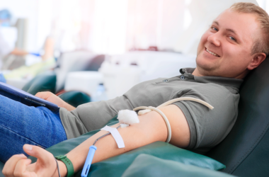 A man smiling while donating blood, reclined on a chair. A medical tube is connected to his arm, representing World Blood Donor Day.