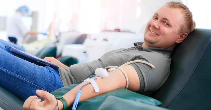 A man smiling while donating blood, reclined on a chair. A medical tube is connected to his arm, representing World Blood Donor Day.