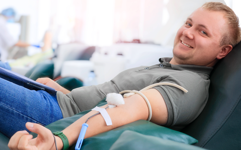 A man smiling while donating blood, reclined on a chair. A medical tube is connected to his arm, representing World Blood Donor Day.