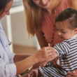 Healthcare provider prepares to vaccinate a young child while a parent offers support.