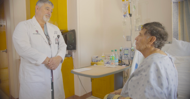 Dr. Philip E. Fiddler, Director of the UMC Burn Center, talks to Javier Diaz, a patient sitting in a hospital bed. The room has medical equipment.