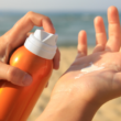 Hands applying sunscreen from an orange bottle on a sandy beach with the ocean in the background, conveying a sense of summer and sun protection.