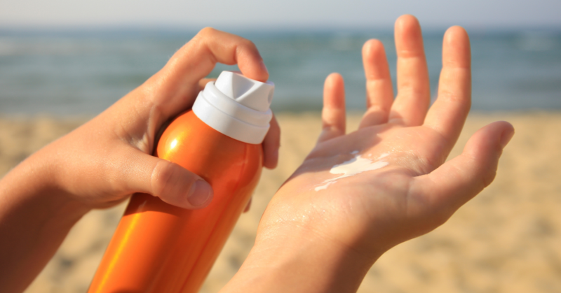 Hands applying sunscreen from an orange bottle on a sandy beach with the ocean in the background, conveying a sense of summer and sun protection.