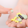 A hand holds various pills: green tablets, yellow round pills, and a black capsule. Another hand picks up a green tablet, conveying a clinical and focused tone.
