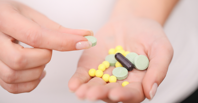 A hand holds various pills: green tablets, yellow round pills, and a black capsule. Another hand picks up a green tablet, conveying a clinical and focused tone.