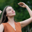 A woman stands outdoors with closed eyes, smiling and raising her arms, enjoying a peaceful moment, representing self-care.