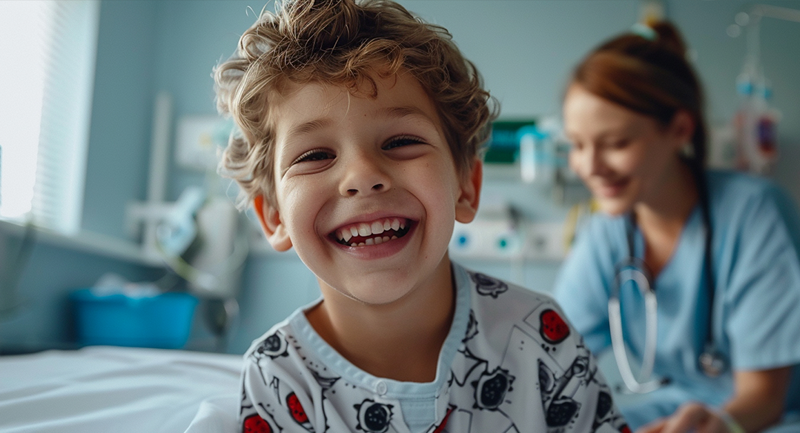 Smiling young child in a hospital gown sits in a pediatric care room with a healthcare professional in the background.