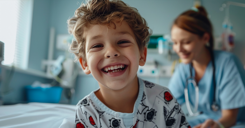 Smiling young child in a hospital gown sits in a pediatric care room with a healthcare professional in the background.