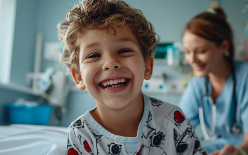 Smiling young child in a hospital gown sits in a pediatric care room with a healthcare professional in the background.