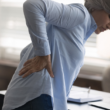 An older woman holds her lower back in discomfort while standing beside a desk.