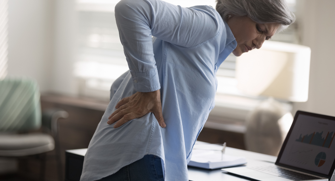 An older woman holds her lower back in discomfort while standing beside a desk.