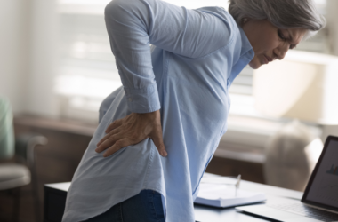 An older woman holds her lower back in discomfort while standing beside a desk.