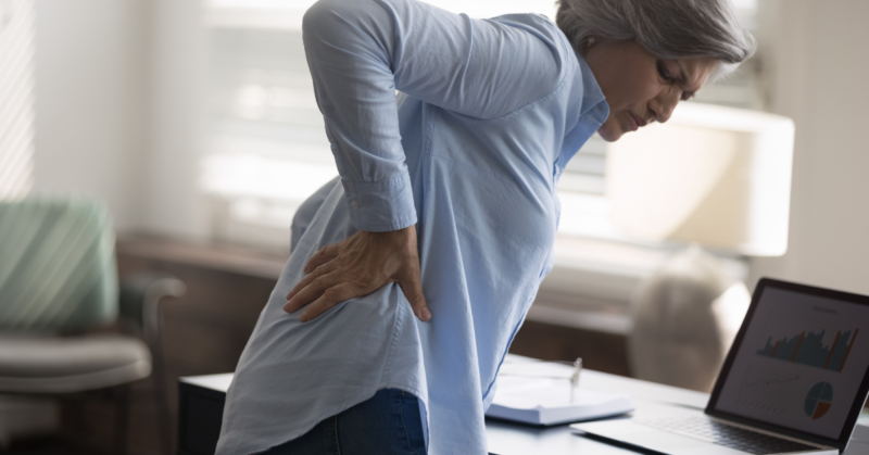 An older woman holds her lower back in discomfort while standing beside a desk.