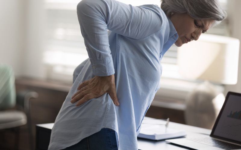 An older woman holds her lower back in discomfort while standing beside a desk.