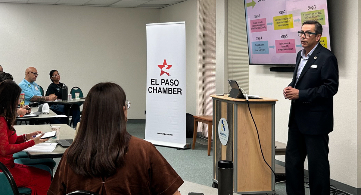 A presenter speaks to attendees during an informational session at the El Paso Chamber, standing beside a screen displaying a step-by-step process while participants listen and take notes.