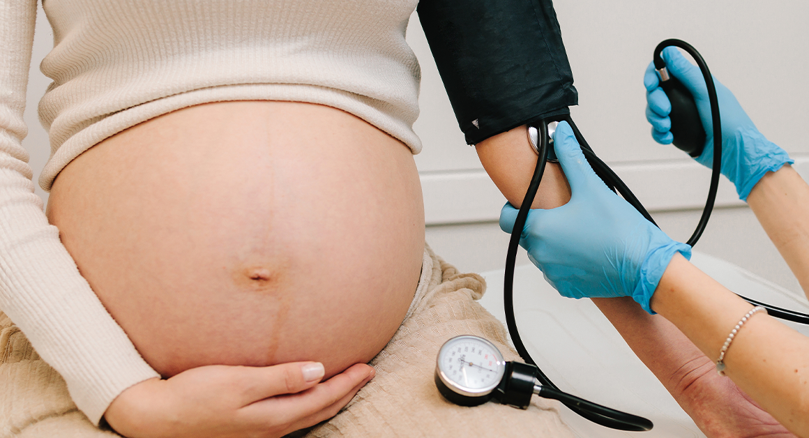 A pregnant woman wearing a white top holds her bare belly. A person wearing blue gloves checks her blood pressure, representing preeclampsia.