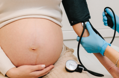 A pregnant woman wearing a white top holds her bare belly. A person wearing blue gloves checks her blood pressure, representing preeclampsia.